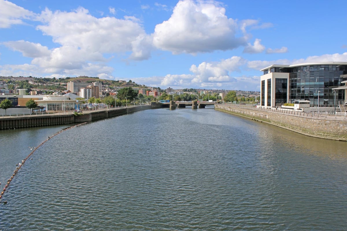 Wide river with modern buildings along both banks, blue sky with white clouds, and hills visible in the distance