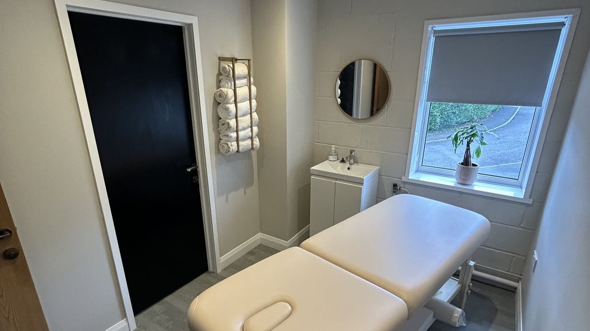 The treatment room at Birchgrove, with a white massage table, a window with a shade drawn and a black door