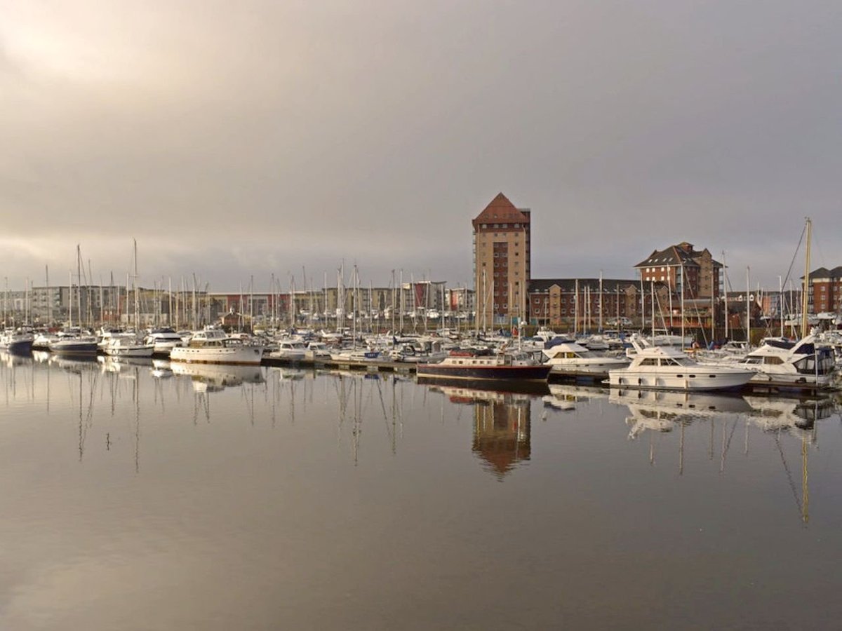 Marina with white sailboats moored in calm water, reflecting historic brick buildings and towers in the background