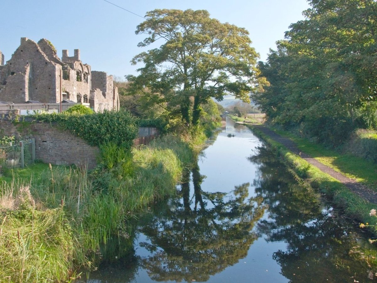 Ruined stone castle beside a calm river lined with green trees and vegetation in summer