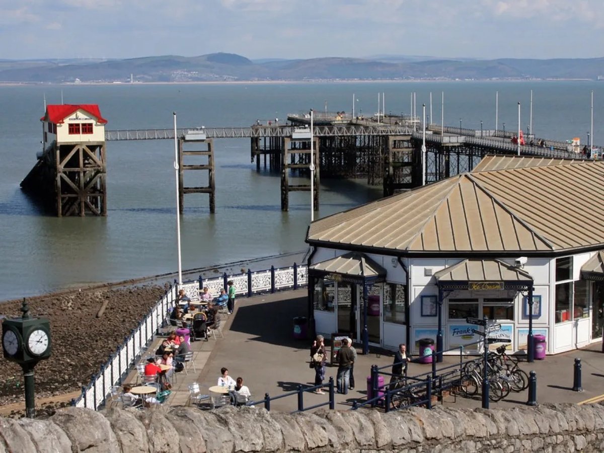 Seaside pier with red lighthouse, wooden docks, and visitor facilities on a clear day with distant hills