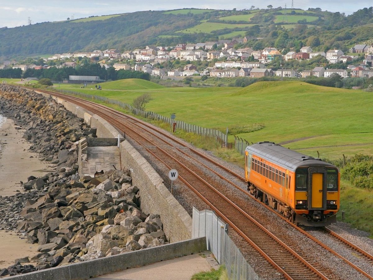 Orange train travelling on railway tracks through green countryside with residential homes on the hillside and a coastal rock barrier in the foreground
