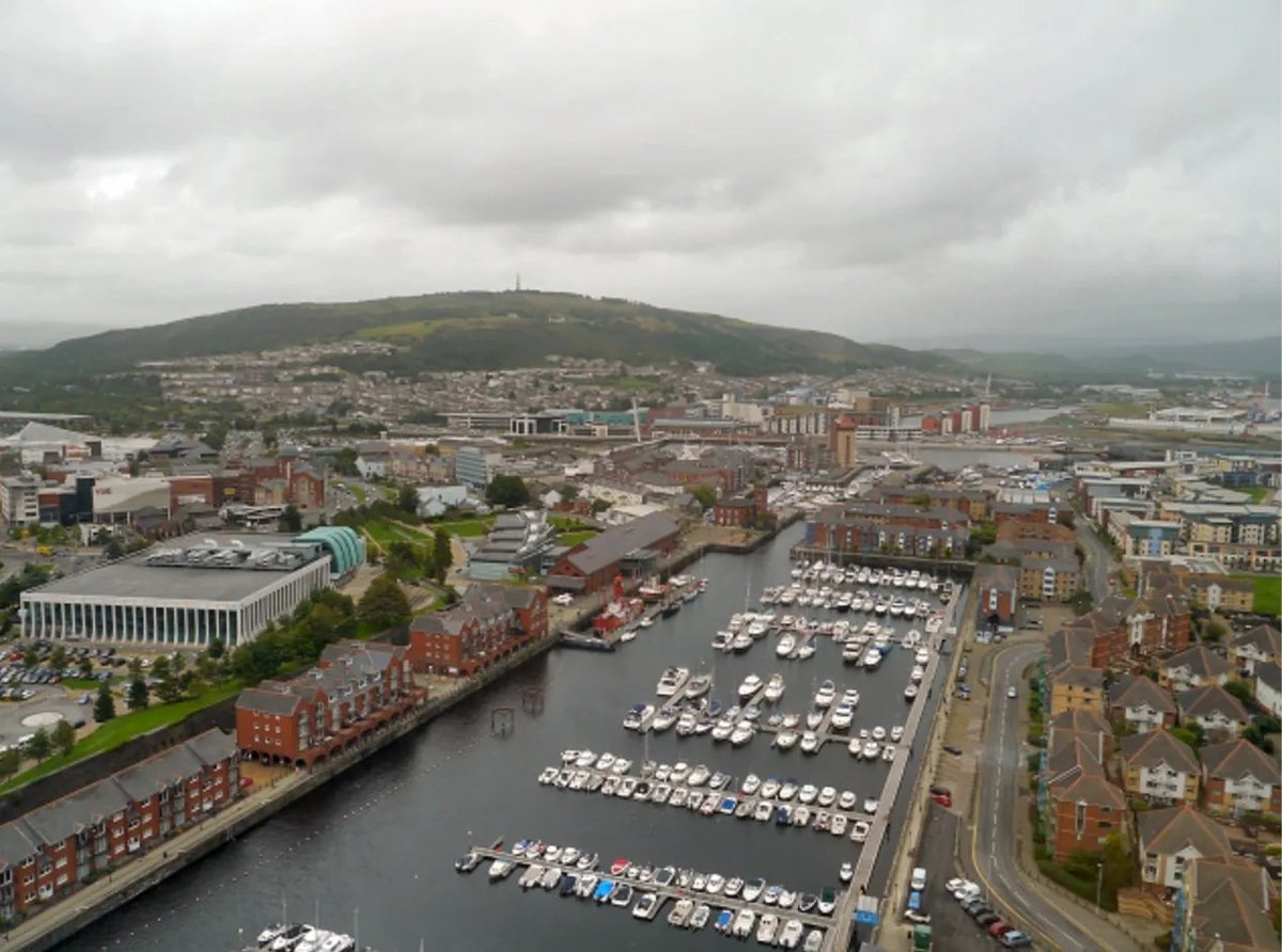 Aerial view of a riverside city with a large marina, brick buildings, and a green mountain in the background under cloudy skies.